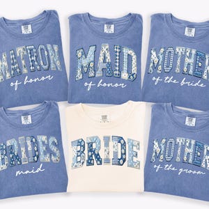 Puede incluir: Seis camisetas en tonos azules y crema. Cada camiseta presenta texto en un dise&ntilde;o de patchwork, con frases como "Matron of Honor", "Maid of Honor", "Mother of the Bride", "Bridesmaid", "Bride" y "Mother of the Groom".