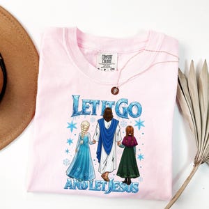May include: A light pink Comfort Colors t-shirt with the text "Let It Go and Let Jesus" in blue and white. The graphic features three figures in a winter scene. A gold necklace with a pendant is on the shirt.