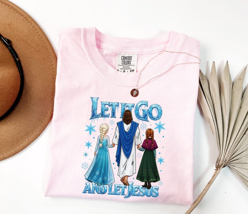May include: A light pink Comfort Colors t-shirt with the text "Let It Go and Let Jesus" in blue and white. The graphic features three figures in a winter scene. A gold necklace with a pendant is on the shirt.