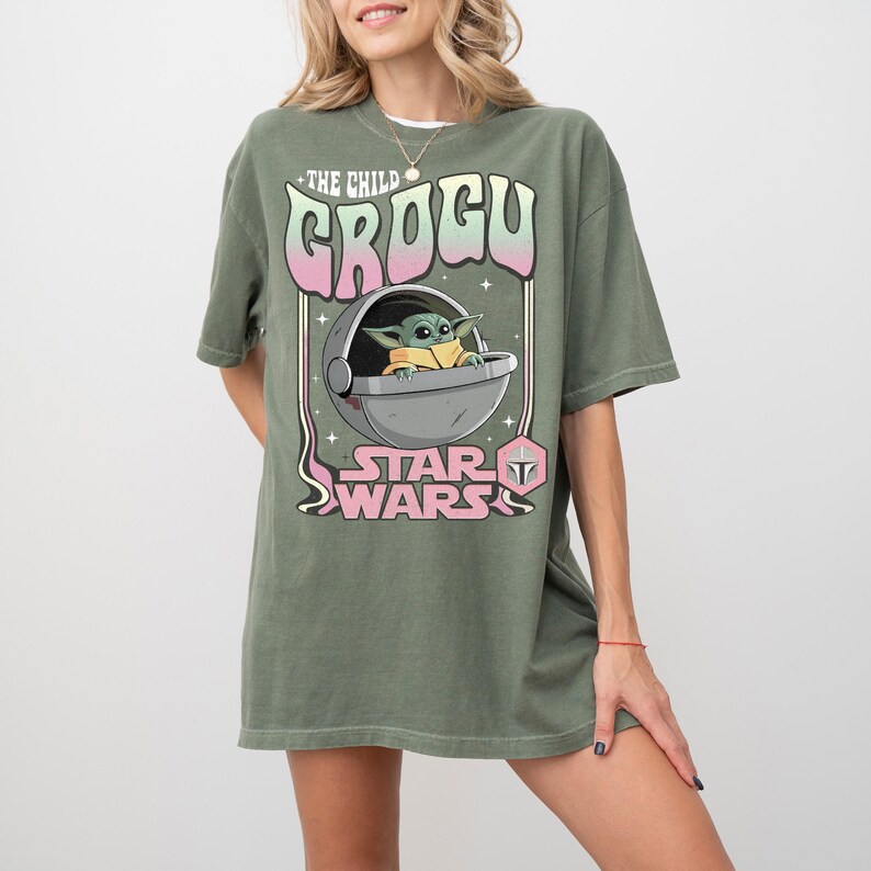 Puede incluir: Una camiseta verde oliva con un estampado gr&aacute;fico del personaje Grogu dentro de una c&aacute;psula flotante. El texto dice THE CHILD GROGU y STAR WARS en una fuente con degradado rosa y blanco. Peque&ntilde;as estrellas rodean la ilustraci&oacute;n central.