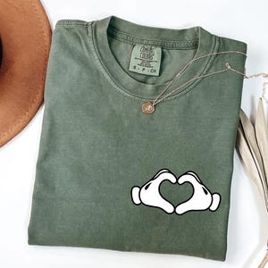 May include: Olive green Comfort Colors t-shirt with a white heart-shaped hand graphic. A gold necklace with a small pendant rests on the shirt. The shirt is folded on a white surface.