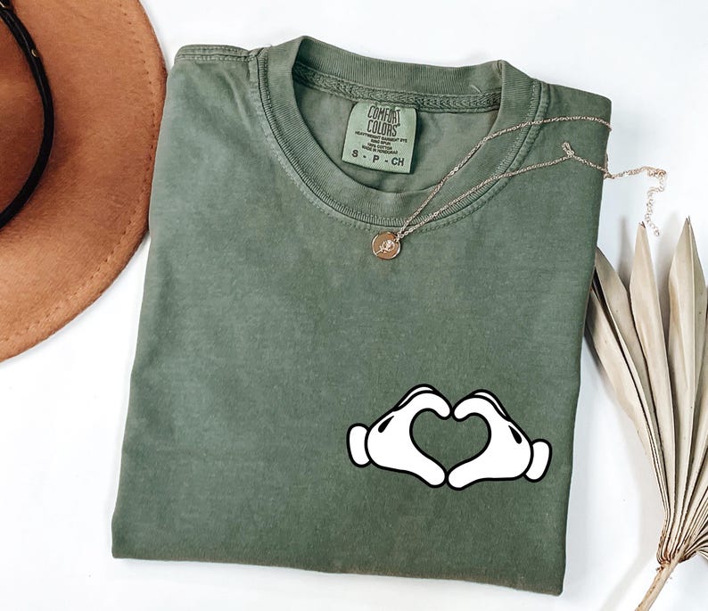 May include: Olive green Comfort Colors t-shirt with a white heart-shaped hand graphic. A gold necklace with a small pendant rests on the shirt. The shirt is folded on a white surface.