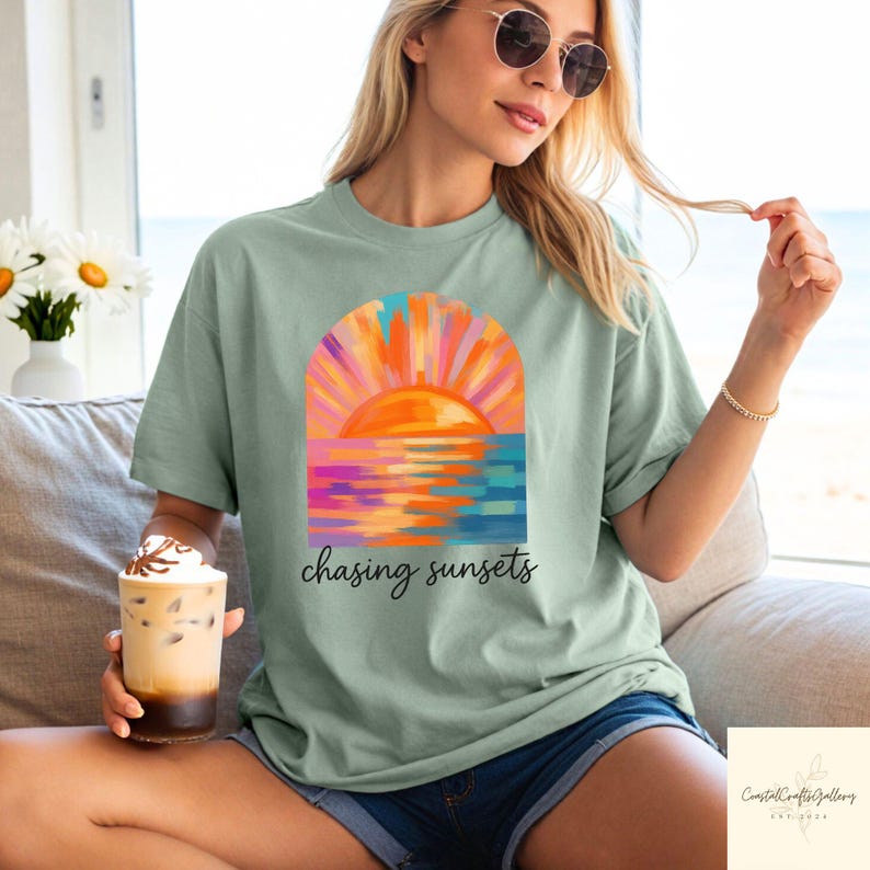 May include: Sage green t-shirt featuring a colorful sunset graphic with the text "chasing sunsets." The sunset design includes shades of orange, pink, and blue. The shirt is a relaxed fit.