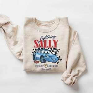 May include: A beige crewneck sweatshirt featuring a blue cartoon car with the text "Lightning Sally" in red and black. The design includes checkered flags and the words "Piston Cup Racing Series 5 Time Champ".