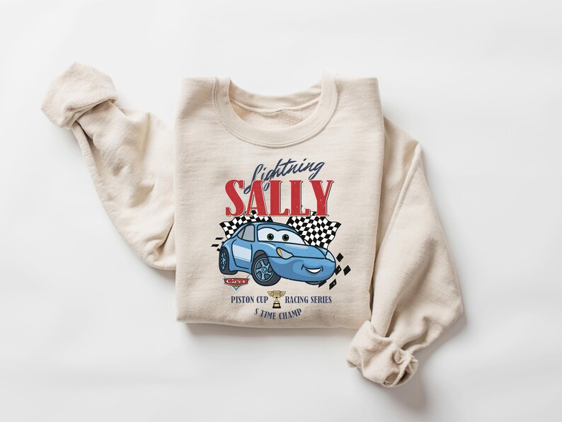 May include: A beige crewneck sweatshirt featuring a blue cartoon car with the text "Lightning Sally" in red and black. The design includes checkered flags and the words "Piston Cup Racing Series 5 Time Champ".