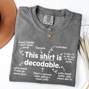 May include: A charcoal gray short-sleeve t-shirt with white text that reads: This shirt is decodable. The graphic includes arrows pointing to linguistic notes like Closed Syllable, Digraphs, r-controlled, Latin-based Prefix, Silent-e, and Latin-based Suffix.