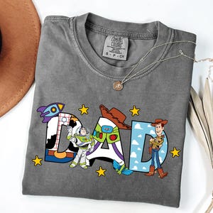 K&ouml;nnte beinhalten: Ein anthrazitfarbenes T-Shirt mit dem Wort DAD in gro&szlig;en Blockbuchstaben. Der Buchstabe D enth&auml;lt eine Weltraum-Ranger-Grafik, der Buchstabe A zeigt einen Cowboyhut und ein Raumanzug-Design, und der Buchstabe D enth&auml;lt eine Cowboy-Figur.