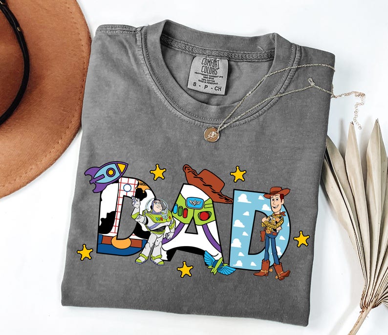 K&ouml;nnte beinhalten: Ein anthrazitfarbenes T-Shirt mit dem Wort DAD in gro&szlig;en Blockbuchstaben. Der Buchstabe D enth&auml;lt eine Weltraum-Ranger-Grafik, der Buchstabe A zeigt einen Cowboyhut und ein Raumanzug-Design, und der Buchstabe D enth&auml;lt eine Cowboy-Figur.