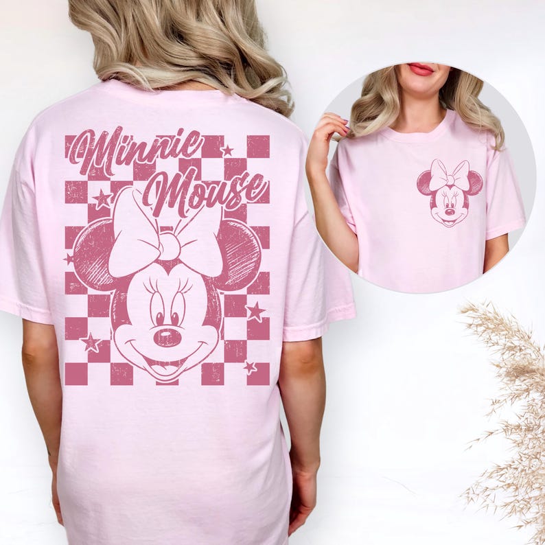 Puede incluir: Camiseta rosa claro con un gr&aacute;fico desgastado de Minnie Mouse. La parte posterior de la camiseta muestra un dise&ntilde;o grande de Minnie Mouse con las palabras "Minnie Mouse" en una fuente de escritura sobre un patr&oacute;n de cuadros. Un gr&aacute;fico m&aacute;s peque&ntilde;o de Minnie Mouse est&aacute; en el frente.
