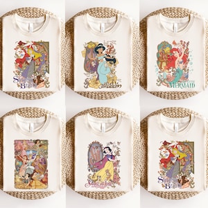 May include: A collection of six cream-coloured t-shirts, each featuring a different graphic print of a classic animated character. The designs include Sleeping Beauty, Aladdin, The Little Mermaid, a group of princesses, and Snow White with floral motifs.