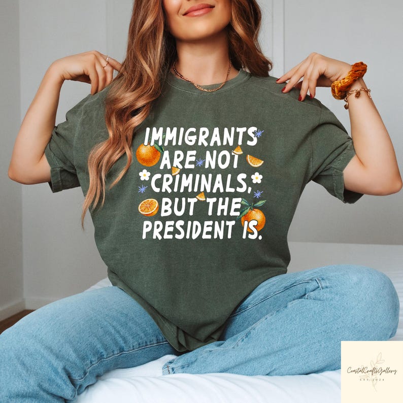 May include: Dark green t-shirt with the text "IMMIGRANTS ARE NOT CRIMINALS, BUT THE PRESIDENT IS." Oranges and floral accents are incorporated into the design. The shirt is a casual wear item.