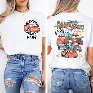 May include: White t-shirt featuring a cartoon car design with the text "NAME" on the front. The back of the shirt displays a colorful graphic of various cartoon cars with the text "Radiator Springs Lightning McQueen".