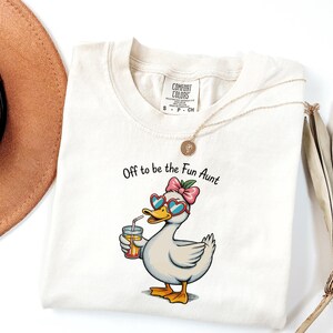 Puede incluir: Una camiseta de color crema con un gráfico de un pato con gafas de sol en forma de corazón y un lazo rosa, sosteniendo un vaso de zumo de naranja. El texto sobre el pato dice, Off to be the Fun Aunt. La camiseta tiene una etiqueta Comfort Colors.