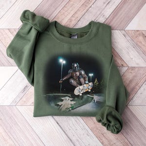 May include: An olive green sweatshirt featuring a graphic of a character from a science fiction series skateboarding. The graphic also includes a small green character on a skateboard. The sweatshirt is laid on a wooden surface.