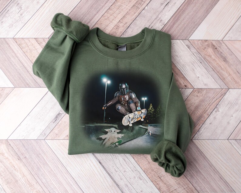 May include: An olive green sweatshirt featuring a graphic of a character from a science fiction series skateboarding. The graphic also includes a small green character on a skateboard. The sweatshirt is laid on a wooden surface.