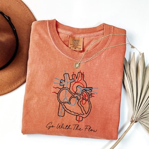 May include: Coral Comfort Colors t-shirt with an anatomical heart illustration in black, red, and blue. The text "Go With The Flow" is printed below the graphic. A gold necklace and a brown hat are also visible.