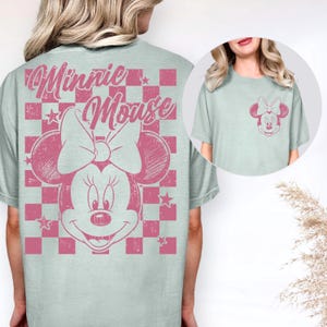Puede incluir: Camiseta azul claro con un dise&ntilde;o rosa de Minnie Mouse. El gr&aacute;fico incluye la cara de Minnie, su lazo y las palabras "Minnie Mouse" en una fuente desgastada. El dise&ntilde;o est&aacute; sobre un fondo a cuadros rosa y blanco. Una versi&oacute;n m&aacute;s peque&ntilde;a del gr&aacute;fico est&aacute; en el bolsillo.