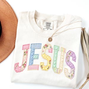 May include: Cream-colored t-shirt with the word "JESUS" appliqued in colorful patchwork fabric. The letters feature floral, gingham, and striped patterns. A gold necklace with a pendant and a brown hat are also in the image.