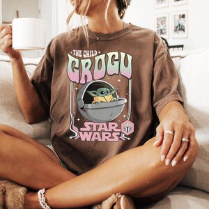 Puede incluir: Una camiseta marr&oacute;n de manga corta con un estampado gr&aacute;fico de Grogu en una c&aacute;psula flotante. El texto dice THE CHILD GROGU STAR WARS en una fuente de estilo retro con colores rosa y verde pastel. El dise&ntilde;o incluye estrellas y un icono de casco.