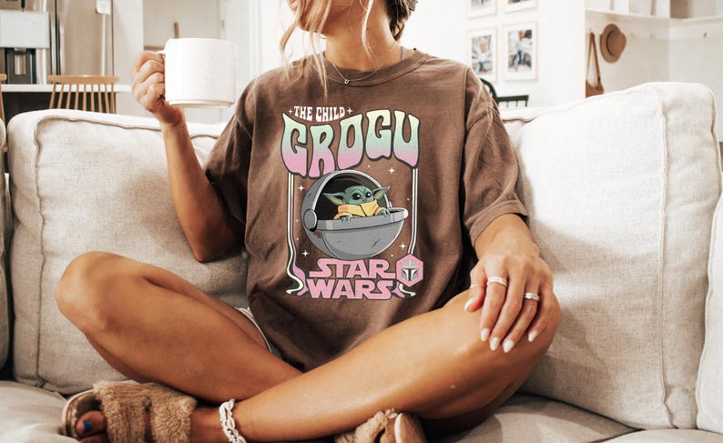 Puede incluir: Una camiseta marr&oacute;n de manga corta con un estampado gr&aacute;fico de Grogu en una c&aacute;psula flotante. El texto dice THE CHILD GROGU STAR WARS en una fuente de estilo retro con colores rosa y verde pastel. El dise&ntilde;o incluye estrellas y un icono de casco.