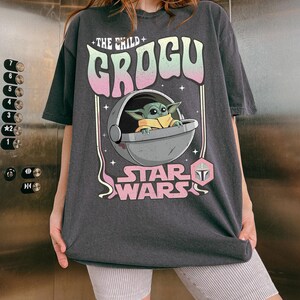 Puede incluir: Una camiseta gris carb&oacute;n de manga corta con un estampado gr&aacute;fico de Grogu en una c&aacute;psula flotante. El texto dice THE CHILD GROGU y STAR WARS en una fuente de degradado pastel. El dise&ntilde;o incluye peque&ntilde;os acentos de estrellas y un icono hexagonal rosa.