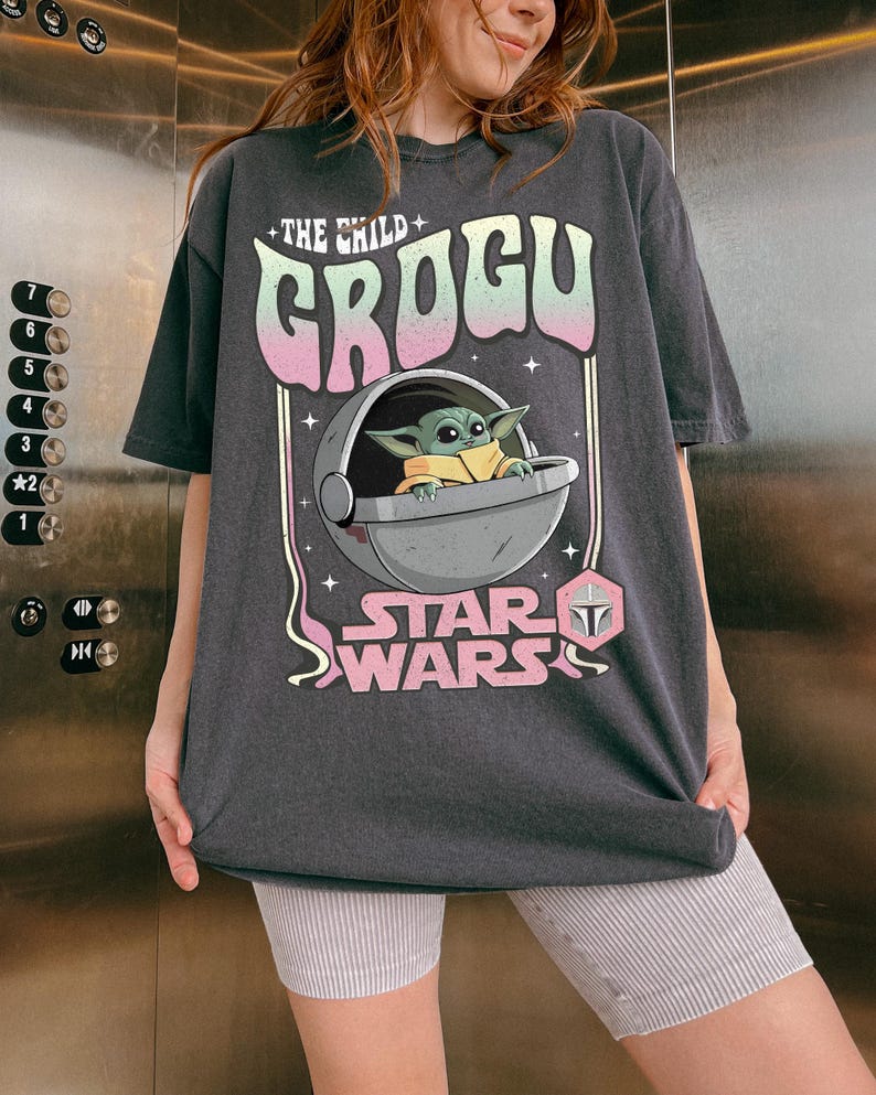 Puede incluir: Una camiseta gris carb&oacute;n de manga corta con un estampado gr&aacute;fico de Grogu en una c&aacute;psula flotante. El texto dice THE CHILD GROGU y STAR WARS en una fuente de degradado pastel. El dise&ntilde;o incluye peque&ntilde;os acentos de estrellas y un icono hexagonal rosa.