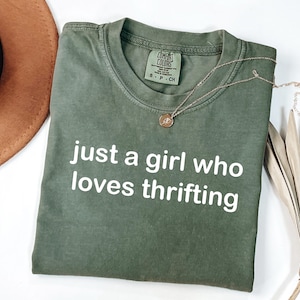 May include: An olive green t-shirt with the text "just a girl who loves thrifting" in white. A gold necklace with a circular pendant rests on the shirt. A brown hat and dried palm leaves are in the background.