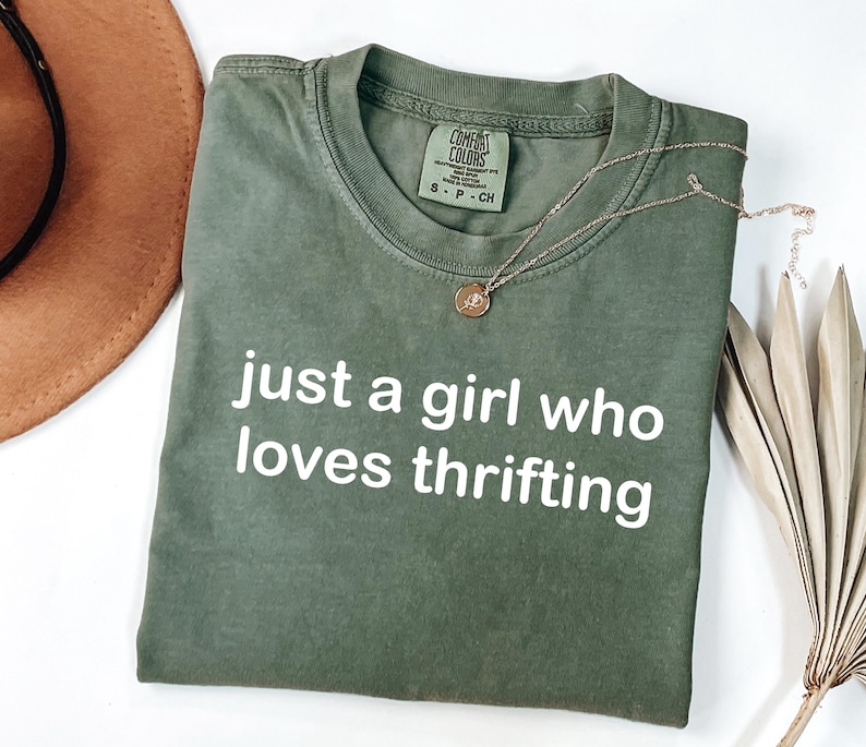 May include: An olive green t-shirt with the text "just a girl who loves thrifting" in white. A gold necklace with a circular pendant rests on the shirt. A brown hat and dried palm leaves are in the background.