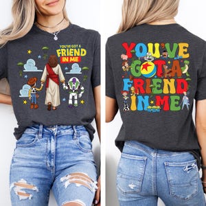 Op de afbeelding: Donkergrijs t-shirt met de tekst "YOU'VE GOT A FRIEND IN ME" en een afbeelding van Jezus, Woody en Buzz Lightyear. De achterkant van het shirt heeft dezelfde tekst in kleurrijke letters met Toy Story-personages.