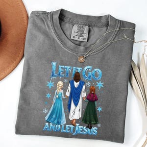 May include: Gray Comfort Colors t-shirt featuring the text "Let It Go and Let Jesus" with an illustration of Jesus and two characters. The shirt has a crew neck and short sleeves. A silver necklace with a pendant is on the shirt.
