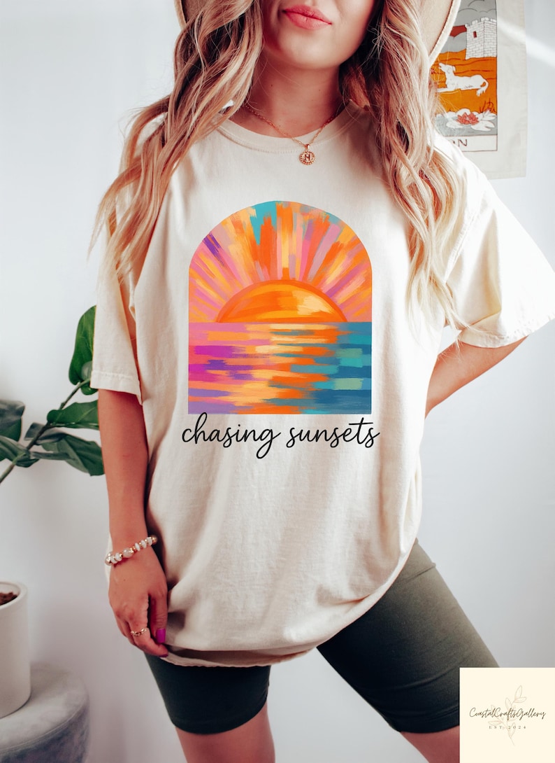 May include: Cream-colored t-shirt featuring a vibrant sunset design with orange, pink, and blue hues. The graphic includes a stylized sun over water, with the words "chasing sunsets" in a script font.