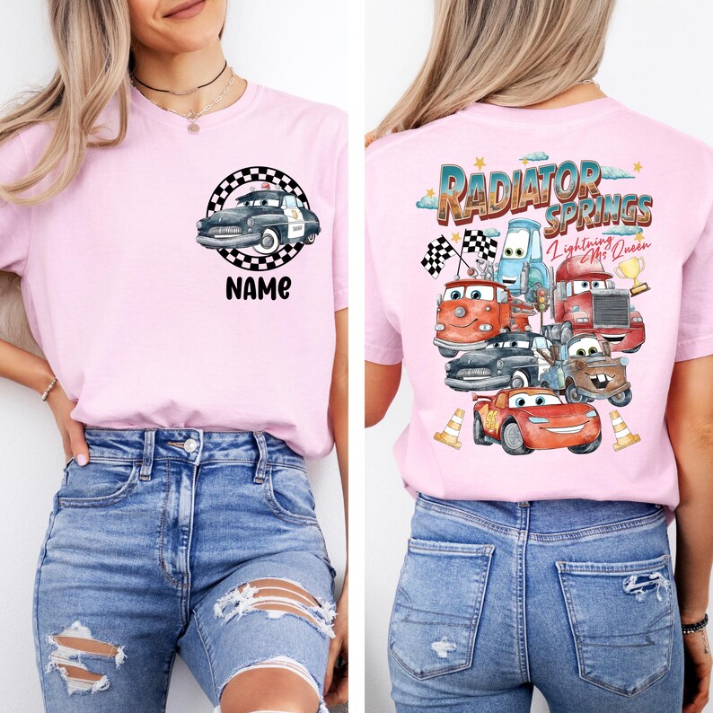 May include: A light pink t-shirt featuring a graphic of a police car inside a checkered circle with the word "NAME" below it. The back of the shirt displays a colorful design of characters from the movie "Cars" with the text "Radiator Springs".