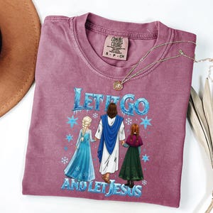 May include: A mauve Comfort Colors t-shirt with the text "LET IT GO AND LET JESUS" in blue. The graphic depicts Jesus holding hands with two animated characters. A gold necklace rests on the shirt.