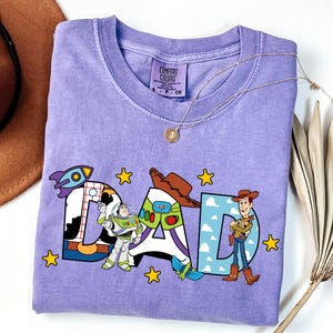 K&ouml;nnte beinhalten: Ein hellviolettes Kurzarm-T-Shirt mit dem Wort DAD in gro&szlig;en Buchstaben. Das D hat ein Kuhfleckenmuster, das A zeigt ein Buzz Lightyear-Design mit einer Rakete und das D enth&auml;lt Woody. Gelbe Sterne umgeben den Schriftzug.