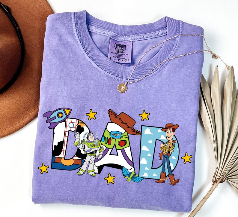 K&ouml;nnte beinhalten: Ein hellviolettes Kurzarm-T-Shirt mit dem Wort DAD in gro&szlig;en Buchstaben. Das D hat ein Kuhfleckenmuster, das A zeigt ein Buzz Lightyear-Design mit einer Rakete und das D enth&auml;lt Woody. Gelbe Sterne umgeben den Schriftzug.