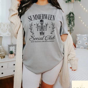 May include: Gray t-shirt with the text "Summerween Social Club" and a skeleton graphic. The shirt also has the phrase "Just Waiting for Halloween". The shirt is paired with white ribbed shorts and a cream-colored cardigan.