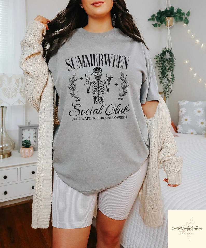 May include: Gray t-shirt with the text "Summerween Social Club" and a skeleton graphic. The shirt also has the phrase "Just Waiting for Halloween". The shirt is paired with white ribbed shorts and a cream-colored cardigan.