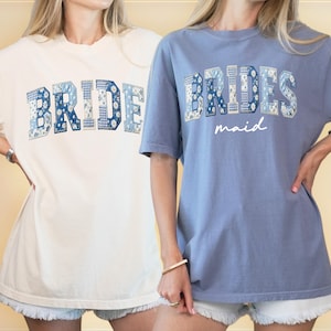 Puede incluir: Dos camisetas de manga corta, una crema y otra azul, con la palabra "BRIDE" en letras de patchwork floral azul. La camisa azul tambi&eacute;n tiene la palabra "maid" en escritura blanca. Ambas camisas son usadas por personas.
