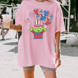May include: Pink t-shirt featuring a cartoon alien wearing a patriotic top hat and holding balloons with Mickey Mouse ears in red, white, and blue. The design includes fireworks and an American flag.