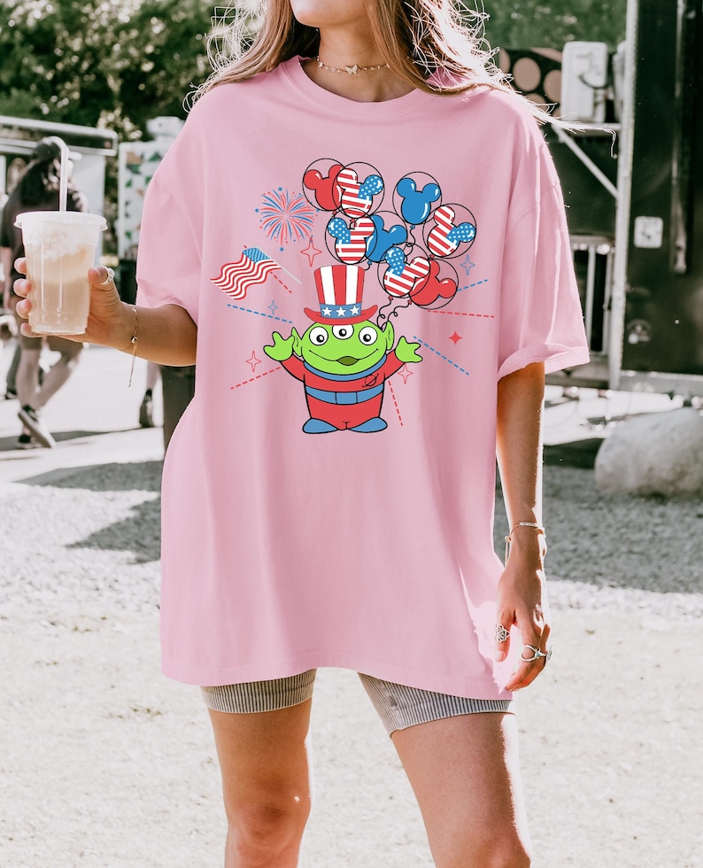 May include: Pink t-shirt featuring a cartoon alien wearing a patriotic top hat and holding balloons with Mickey Mouse ears in red, white, and blue. The design includes fireworks and an American flag.