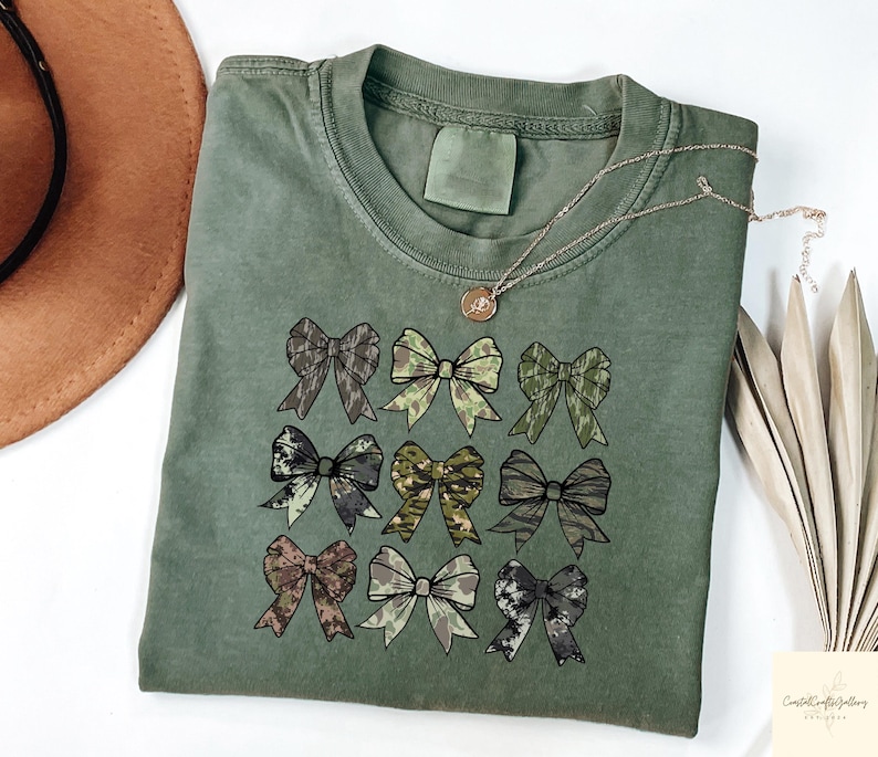 May include: An olive green t-shirt featuring nine bow designs in various camouflage patterns. A gold necklace with a pendant rests on the shirt. The shirt is folded, with a brown hat and dried plant stems in the background.