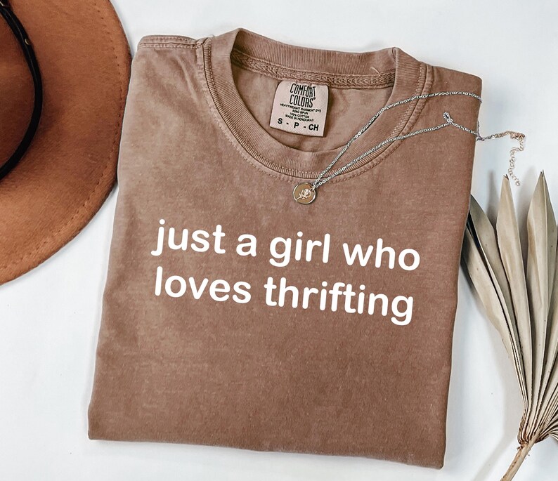May include: A brown t-shirt with the text "just a girl who loves thrifting" in white. A silver necklace with a pendant is draped across the shirt. A brown hat and dried plant are in the background.