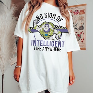 May include: White t-shirt with a Buzz Lightyear graphic and the text "NO SIGN OF INTELLIGENT LIFE ANYWHERE". The design shows Buzz in his spacesuit with green, purple, and blue details. The shirt has a relaxed, oversized fit.
