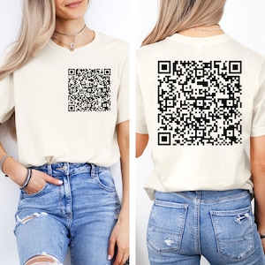 May include: A cream-coloured short-sleeve t-shirt featuring a black QR code graphic on the front left chest and a significantly larger black QR code graphic printed on the back of the garment, paired with blue denim jeans.