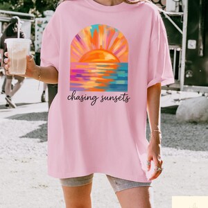 May include: A light pink t-shirt featuring a colorful sunset design with the words "chasing sunsets" in a script font. The sunset art includes orange, yellow, blue, and purple hues. The shirt is a casual, oversized style.