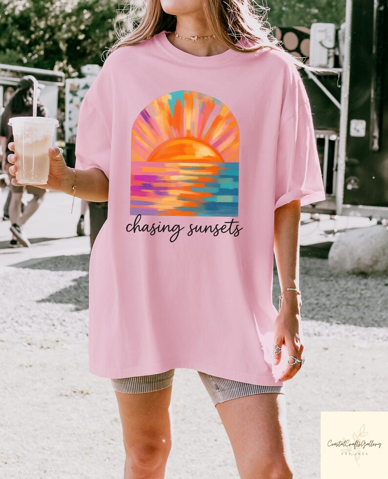 May include: A light pink t-shirt featuring a colorful sunset design with the words "chasing sunsets" in a script font. The sunset art includes orange, yellow, blue, and purple hues. The shirt is a casual, oversized style.