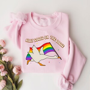 May include: Pink sweatshirt featuring a white goose flying with a rainbow pride flag and the text "Silly Goose on the Loose".