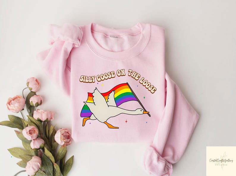 May include: Pink sweatshirt featuring a white goose flying with a rainbow pride flag and the text "Silly Goose on the Loose".