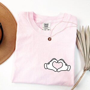 May include: A light pink Comfort Colors t-shirt with a crew neck. The shirt features a graphic of two white cartoon hands forming a heart shape. A gold necklace with a small pendant is on the shirt.