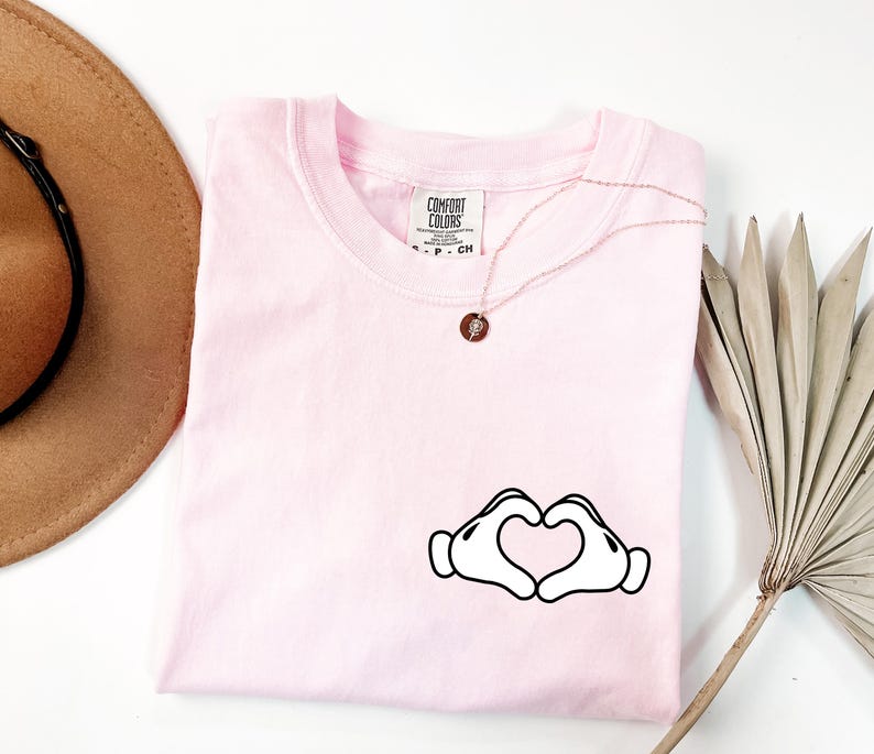 May include: A light pink Comfort Colors t-shirt with a crew neck. The shirt features a graphic of two white cartoon hands forming a heart shape. A gold necklace with a small pendant is on the shirt.